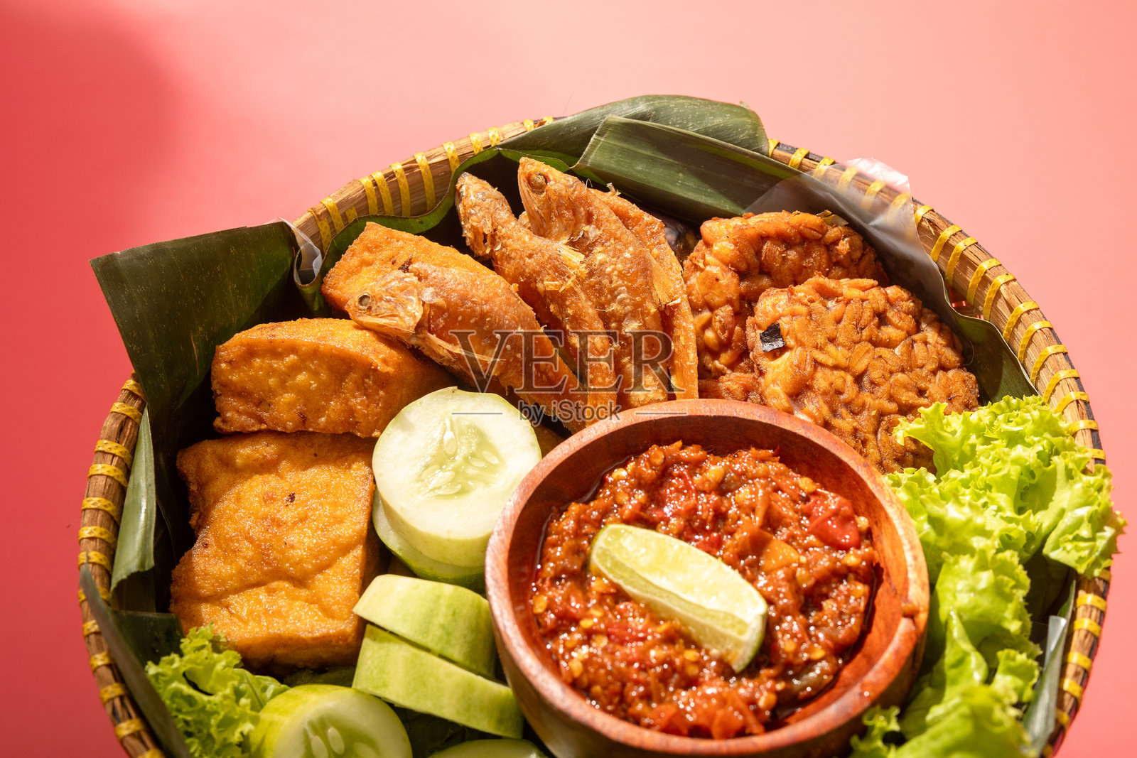 一份传统的印尼餐，盛放在编织的竹篮中，包含炸鱼、豆腐、黄瓜片、生菜和辣椒酱，摆放在香蕉叶上，呈现出色彩鲜艳、令人垂涎的效果。照片摄影图片