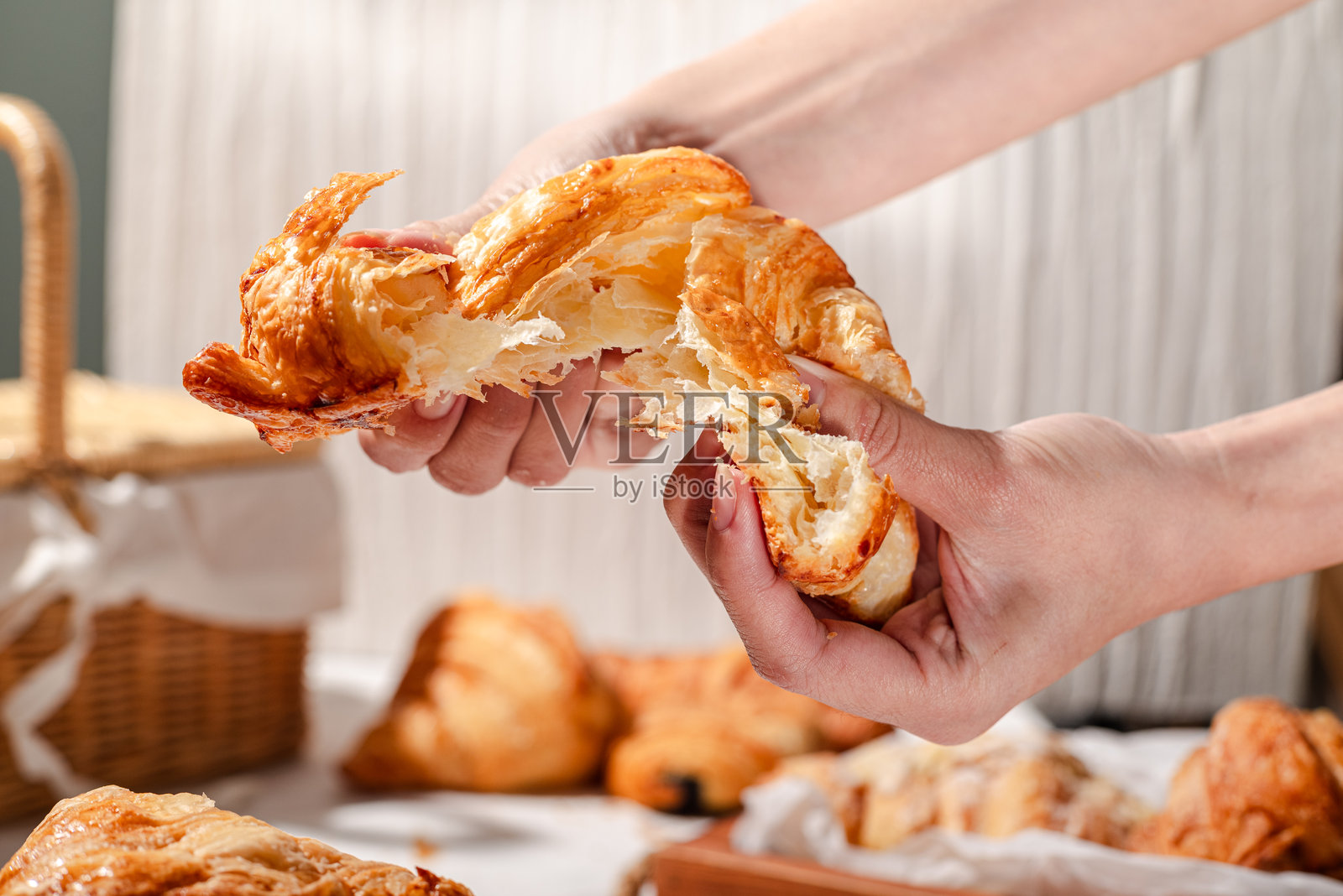 特写镜头展示双手撕开一只新鲜的金色可颂，露出其柔软、轻盈的层次和酥脆的外壳，背景是一个乡村风格的面包店，摆满了糕点、篮子和木质餐盘。照片摄影图片