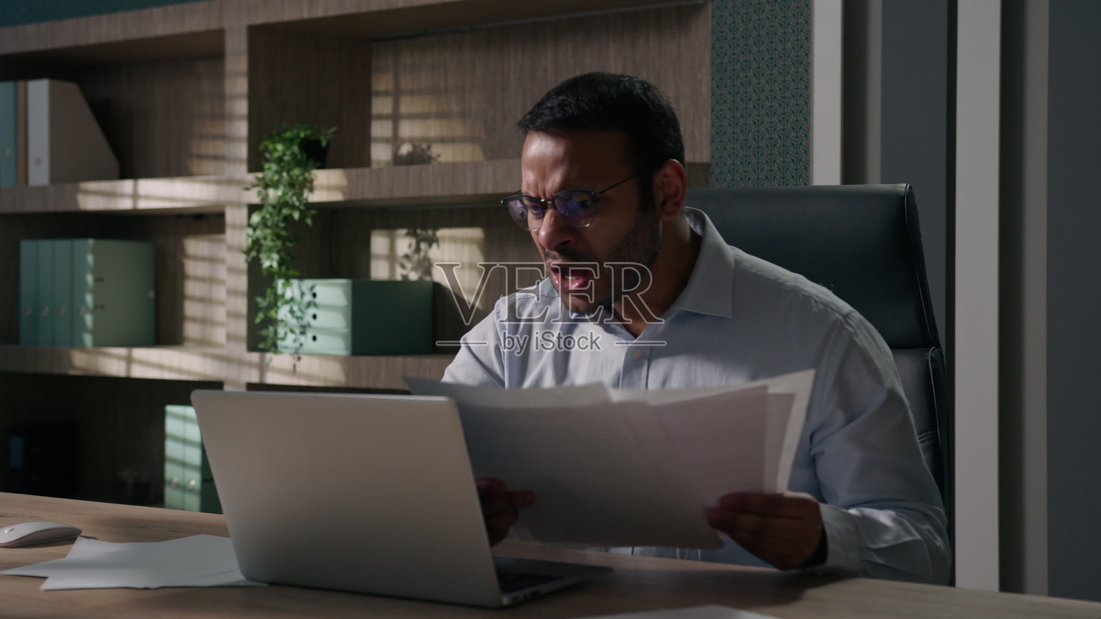 愤怒的印度阿拉伯美国商人感到愤怒、焦虑和攻击性，手中拿着账单，男性公司会计因疲惫而苦恼，愤怒地将文件扔掉，盯着笔记本电脑。照片摄影图片