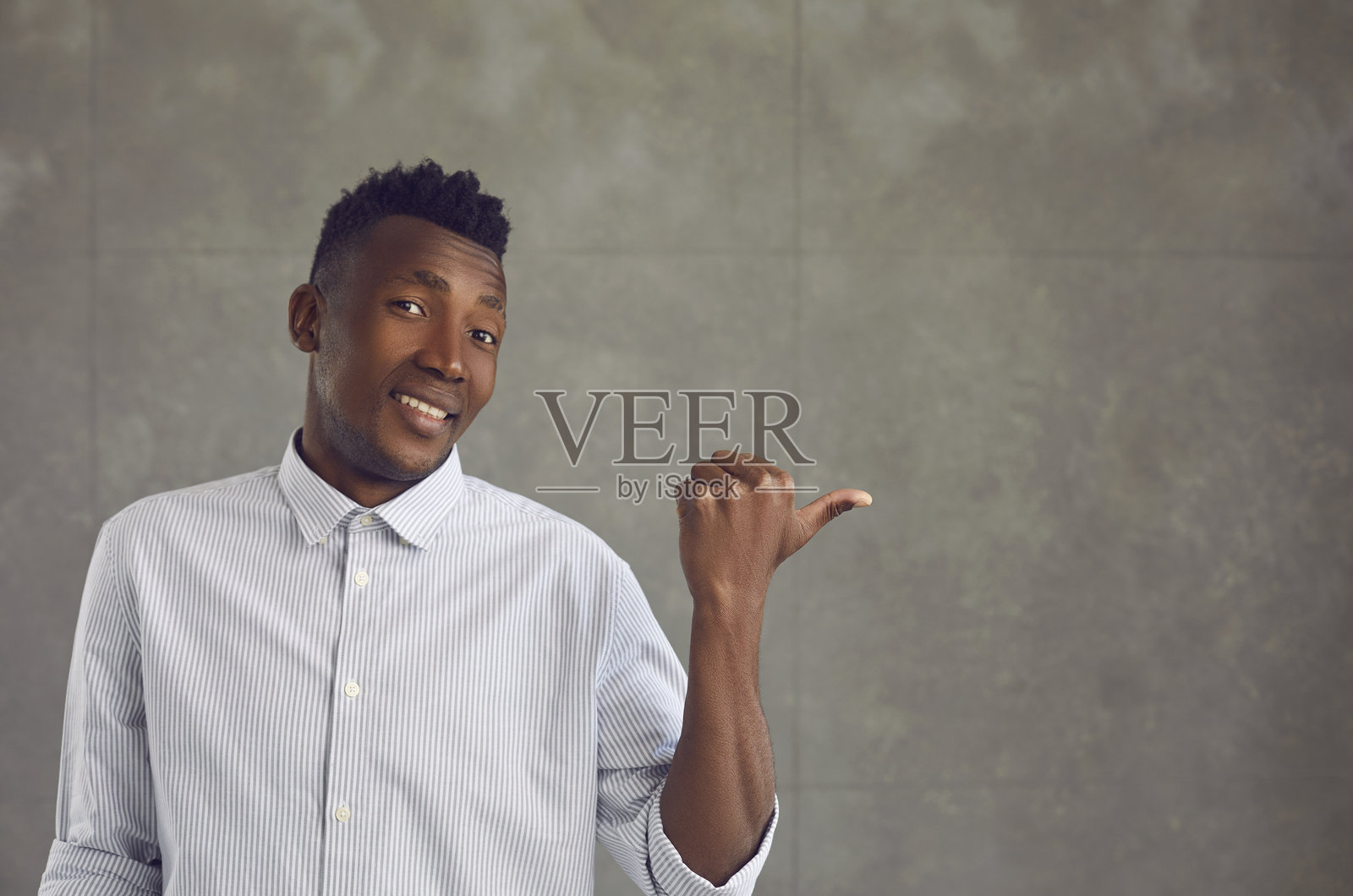 快乐的年轻黑人男子在灰色混凝土墙上用拇指指向广告空白处照片摄影图片