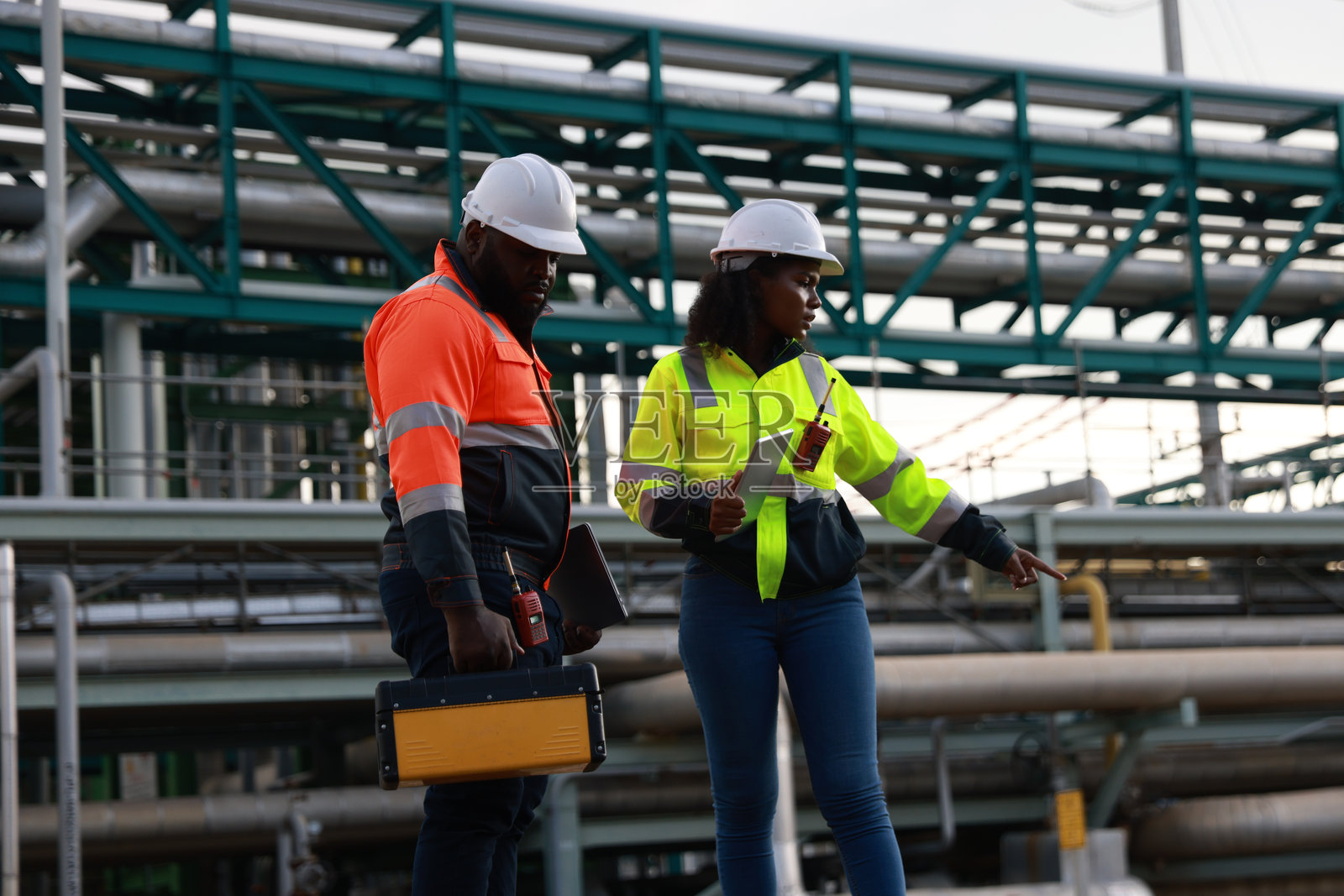 系统工程团队成员对工厂的管道进行了预防性维护，并检查了铁路工作站的调查，背景是石油炼油厂。照片摄影图片