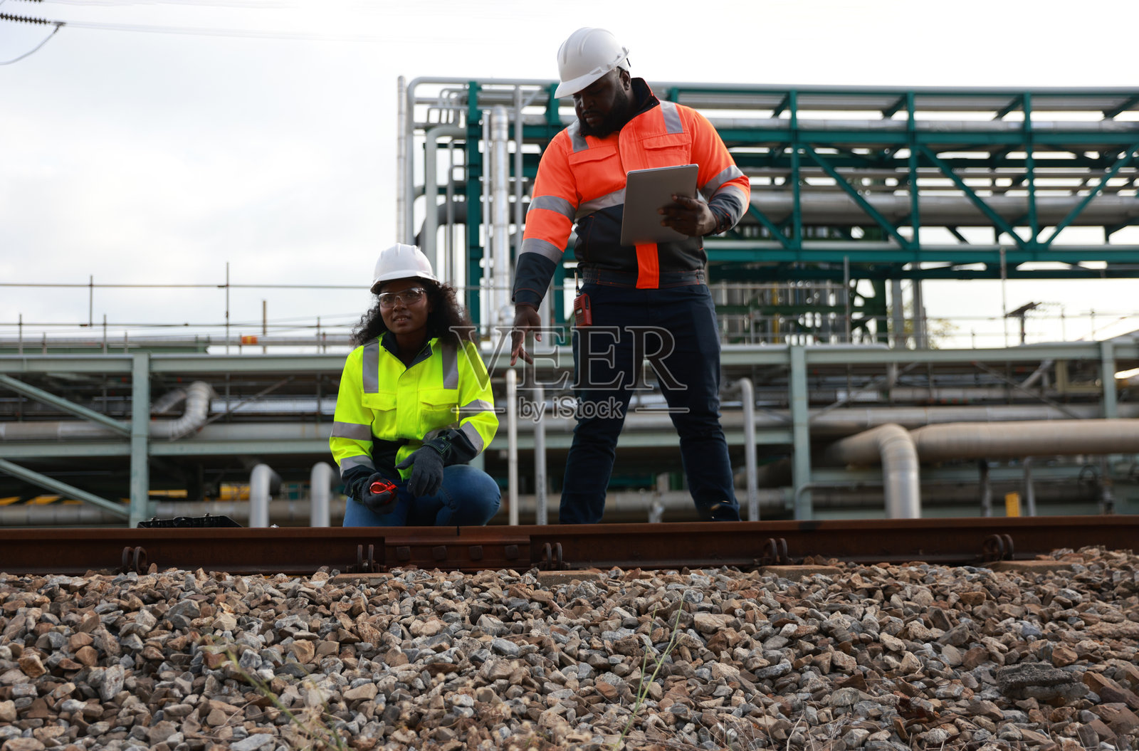 系统工程团队成员对工厂的管道进行了预防性维护，并检查了铁路工作站的调查，背景是石油炼油厂。照片摄影图片