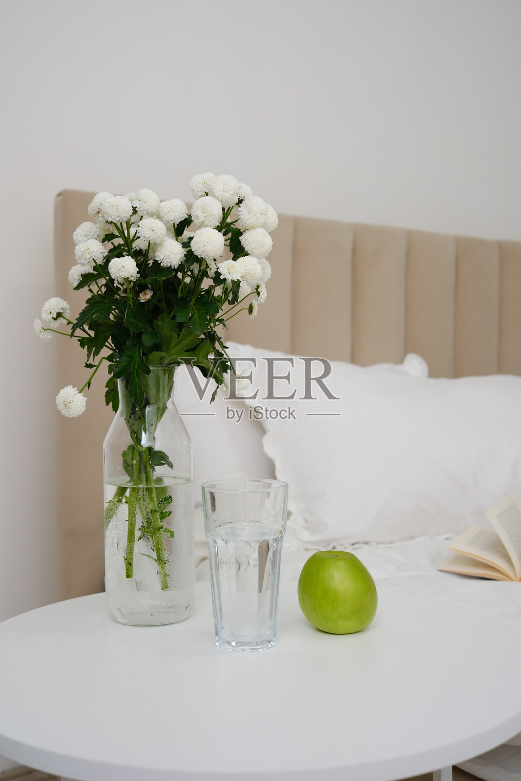 一个宁静、安详、极简的卧室场景，白色圆桌上摆放着花朵、光滑的玻璃和一个鲜艳的绿色苹果。照片摄影图片