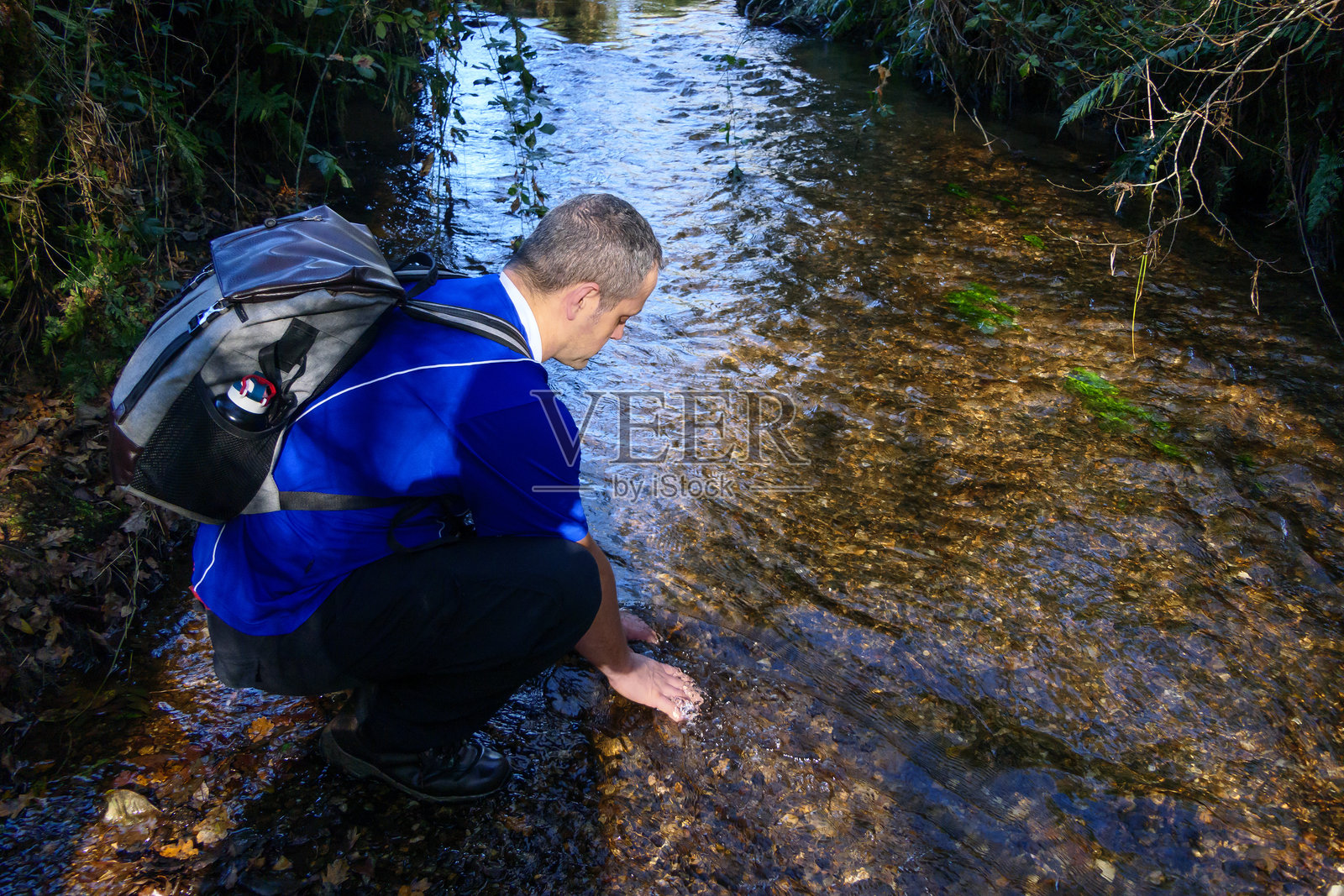 一个男人蹲在河岸上，双手触碰着水面。他背着一个装有水壶的背包。照片摄影图片