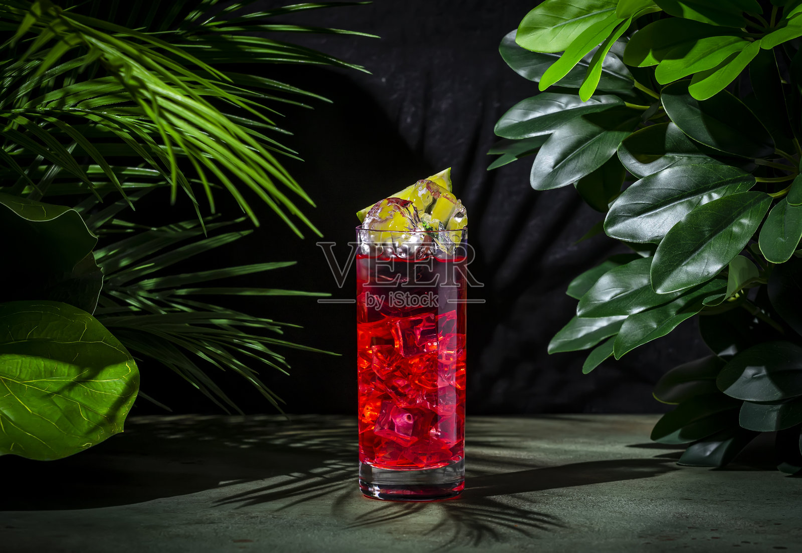 恶魔鸡尾酒，含有龙舌兰酒、黑加仑利口酒、青柠汁、姜啤酒和冰块。深绿色背景配有热带叶子。照片摄影图片