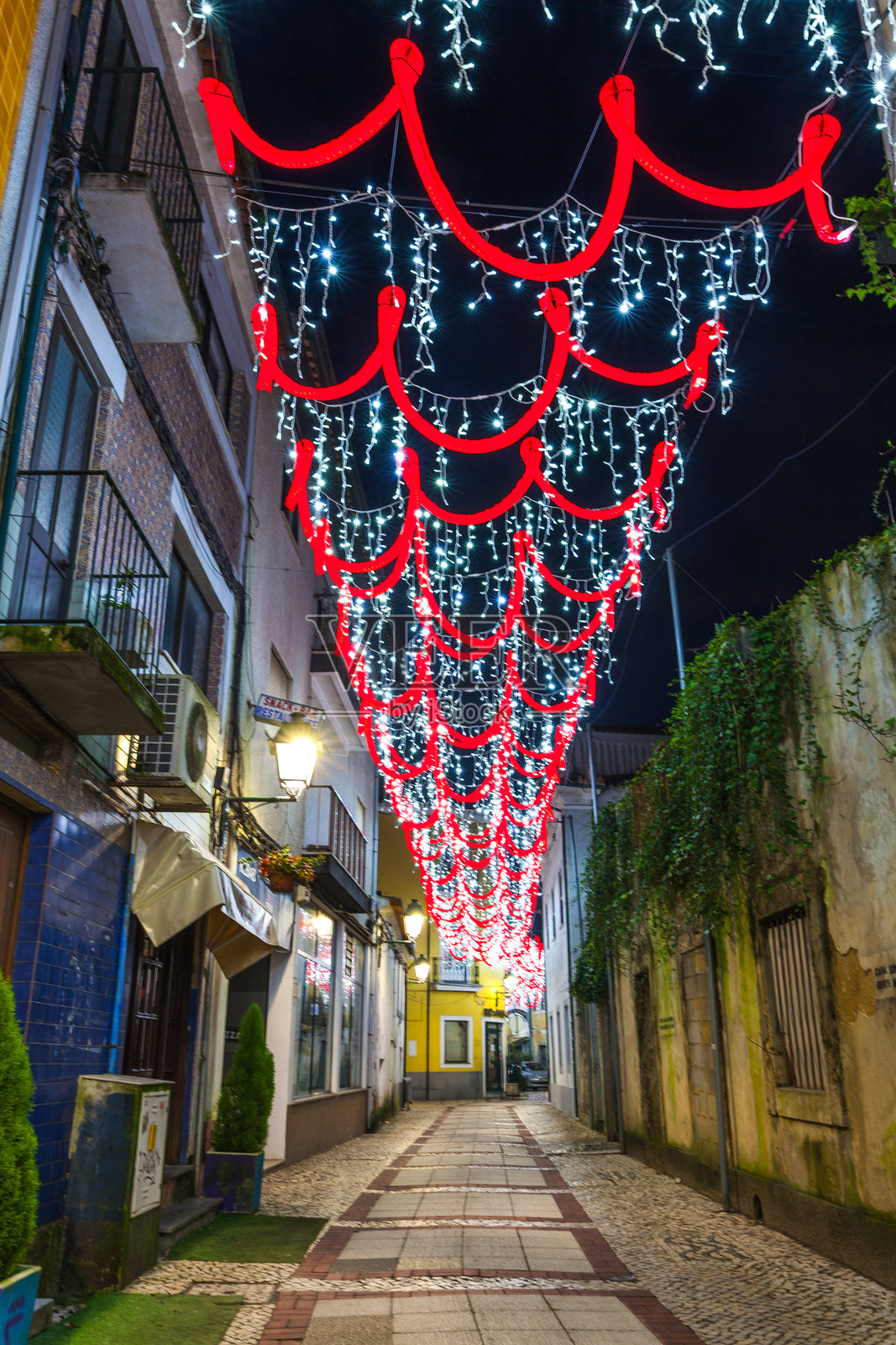 一条狭窄的街道装饰着红色丝带和白色圣诞灯，营造出夜晚的节日气氛。照片摄影图片
