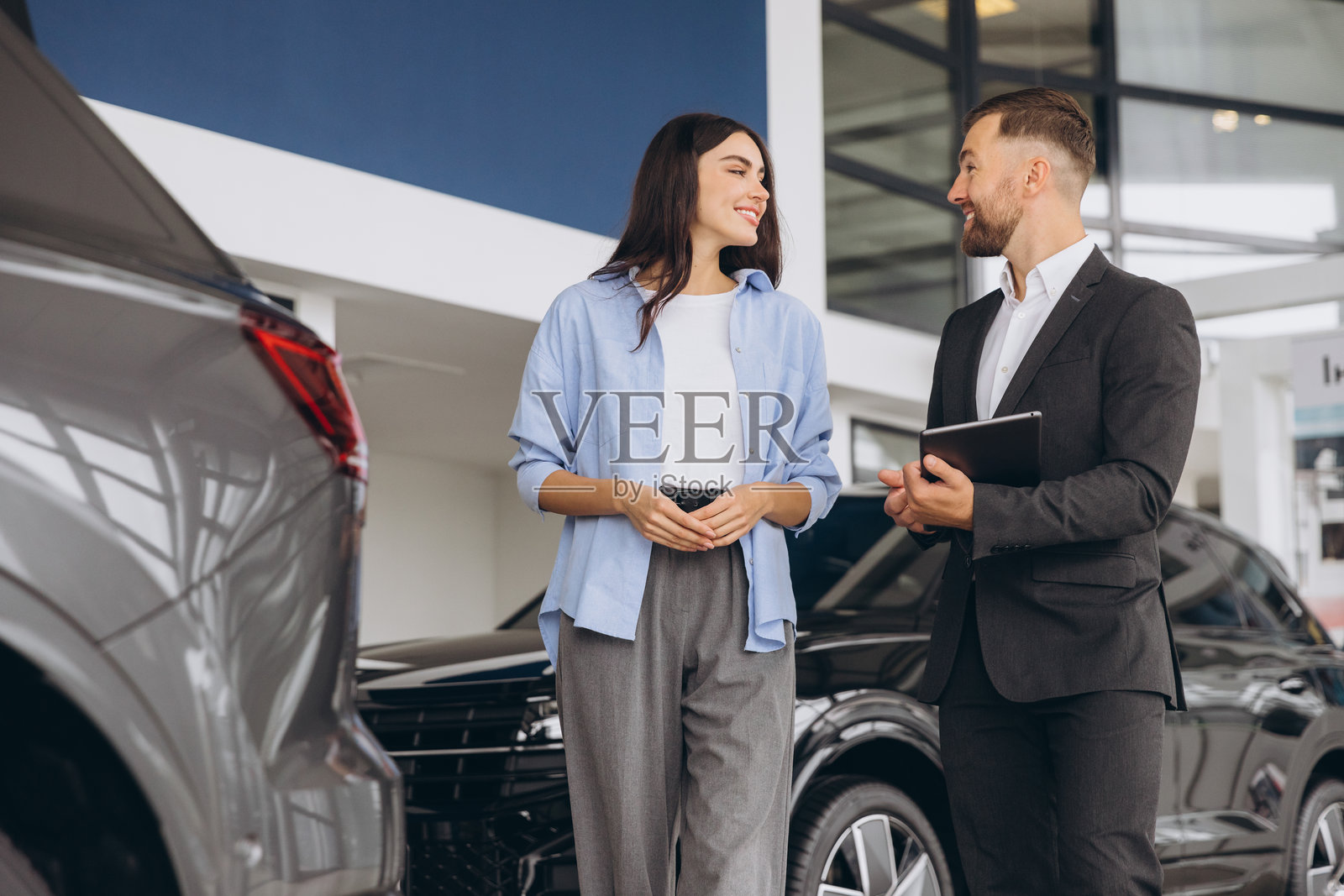 年轻女性顾客穿着衬衫，与销售员咨询，手持平板电脑，选择汽车，想要在汽车展厅购买新车。照片摄影图片