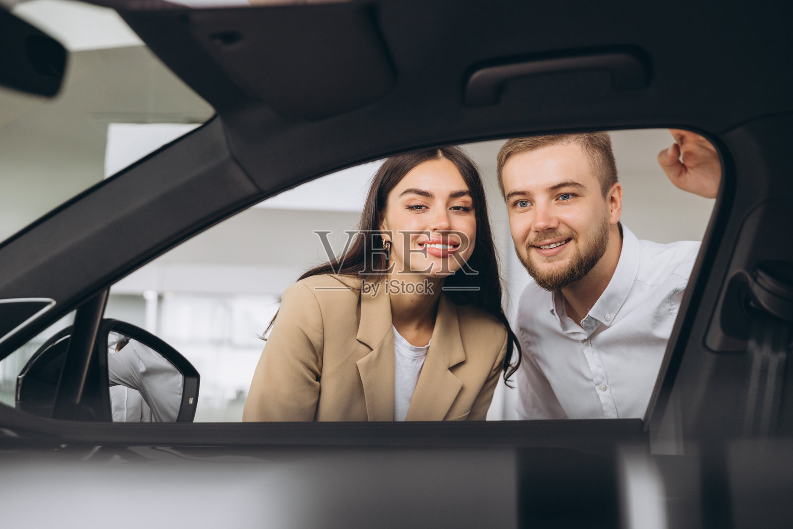 快乐的年轻男女在购买新车，通过窗户查看汽车展厅，全景。微笑的白人夫妇在展厅内查看汽车，准备购买车辆。照片摄影图片
