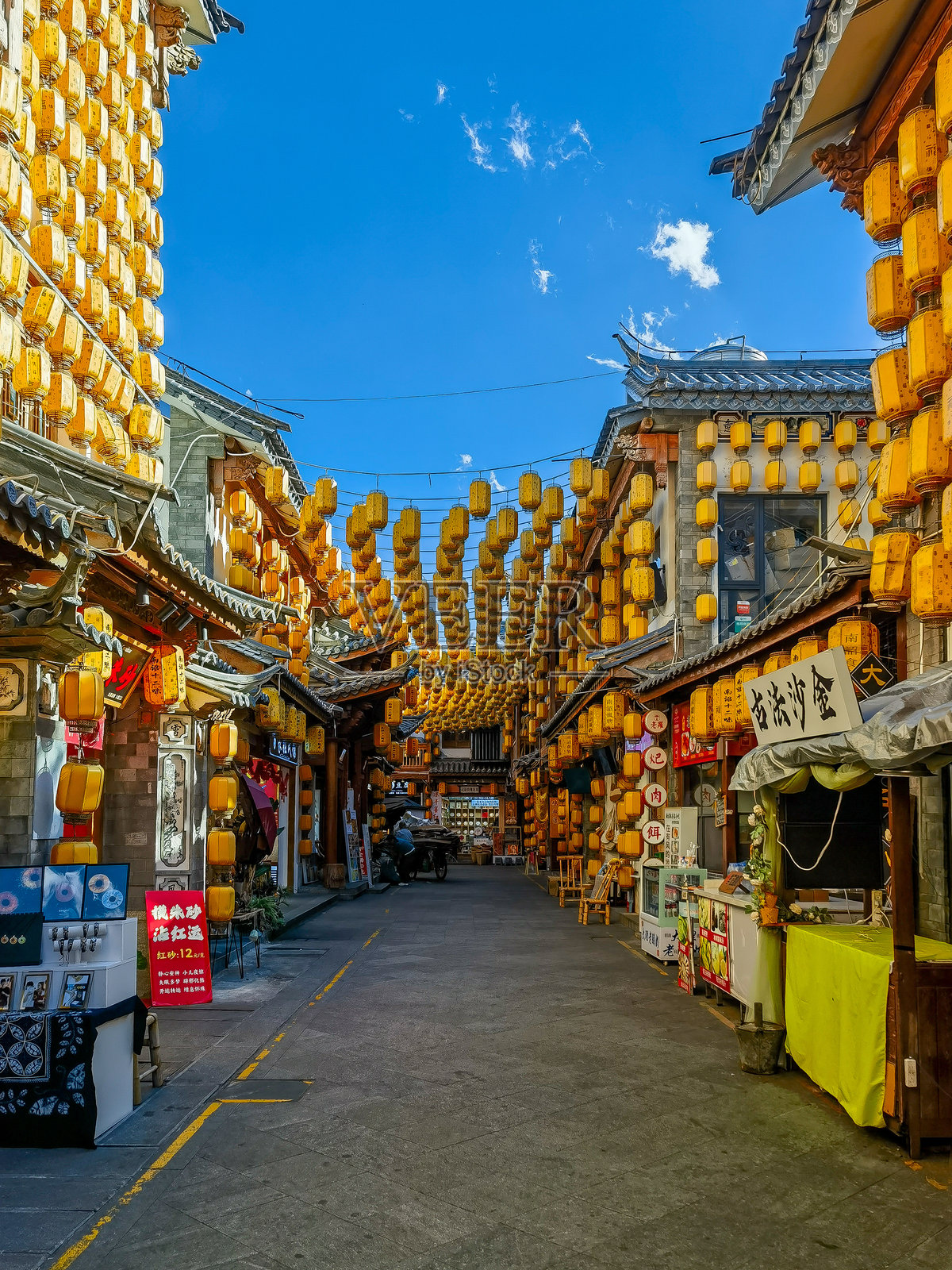 一个充满活力的街景，传统建筑之间挂满了许多黄色灯笼。小路两旁是商店和摊位，营造出在明亮蓝天下热闹的节日气氛。照片摄影图片