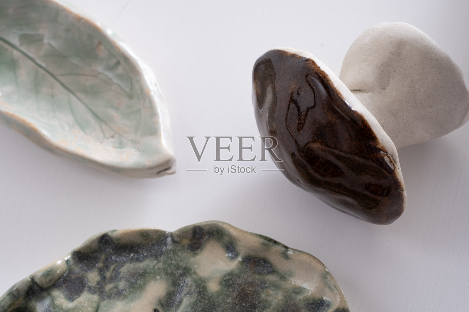 白色表面上的陶瓷装饰特写。包括一个光滑的绿色叶子形状的盘子，一个有纹理的蘑菇雕塑，以及一个带有植物印记的抽象绿色盘子。照片摄影图片