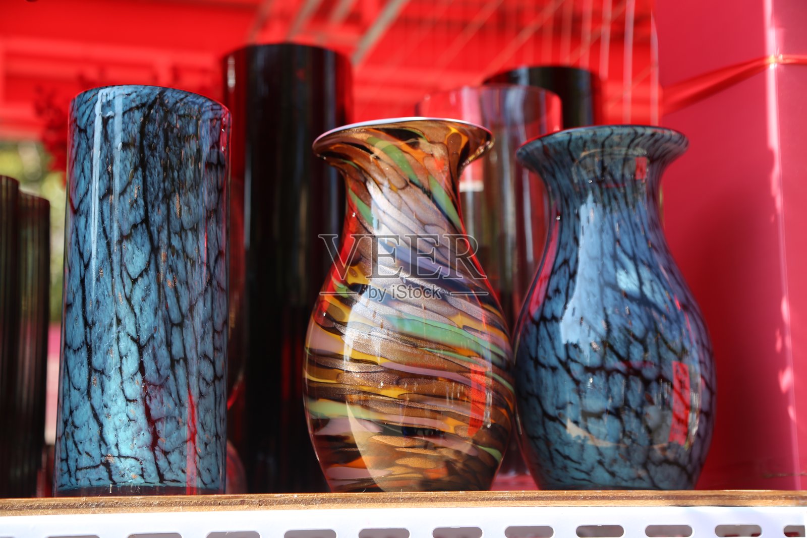 三个不同形状和颜色的美丽玻璃花瓶在粉色墙壁的户外市场摊位上展示。照片摄影图片