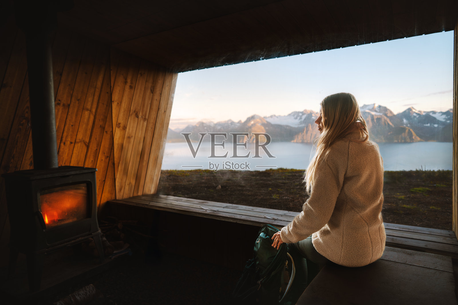 女人在挪威的山间小屋里，透过全景窗户享受壁炉的温暖，旅行生活方式，秋季假期背包旅行，独自在温馨的房子里休息的女徒步旅行者照片摄影图片