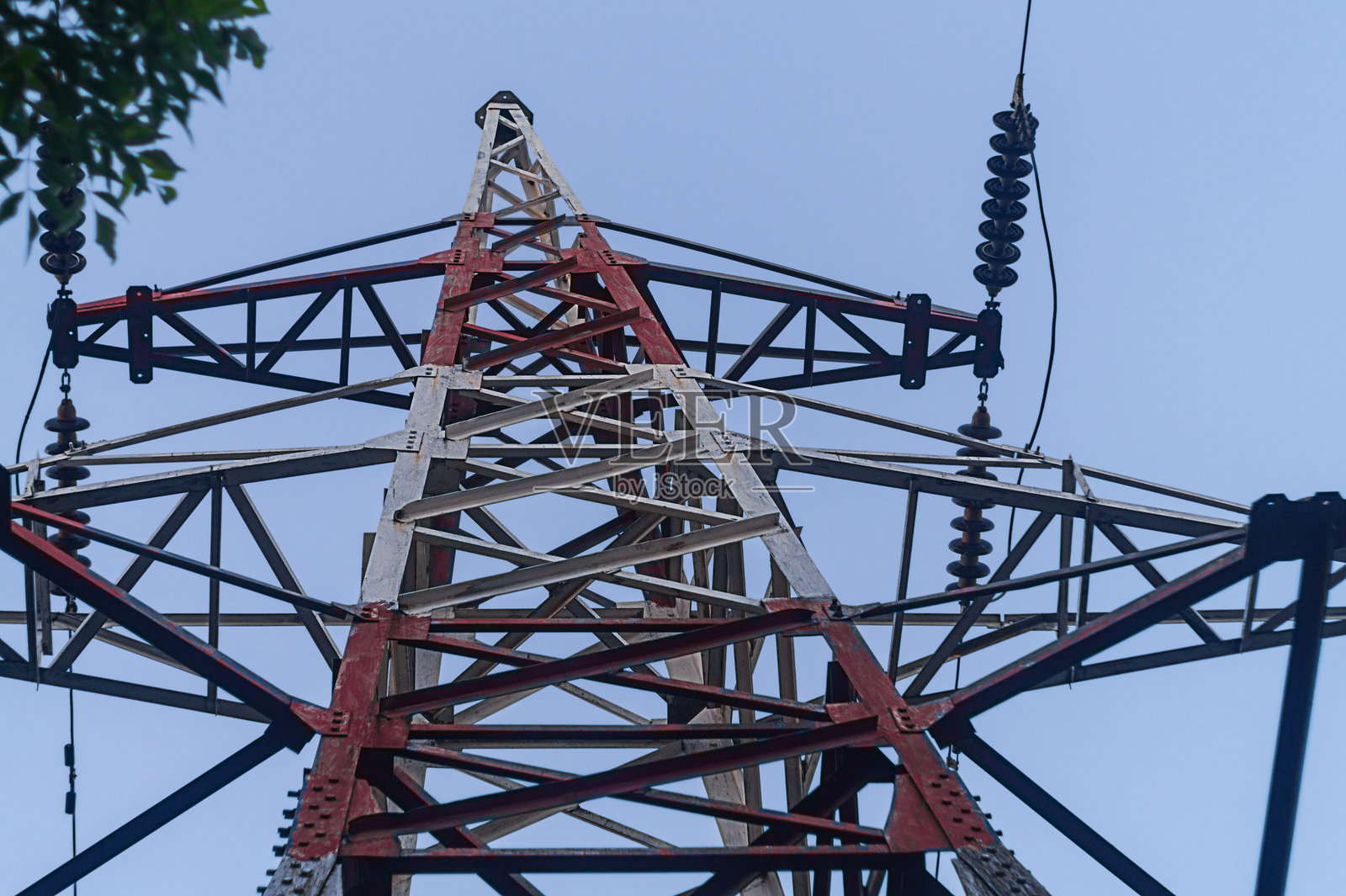 高压电力传输的大型金属杆，红白相间的颜色，背景是蓝天和树枝。底部视角。电力供应基础设施，工业设备。照片摄影图片