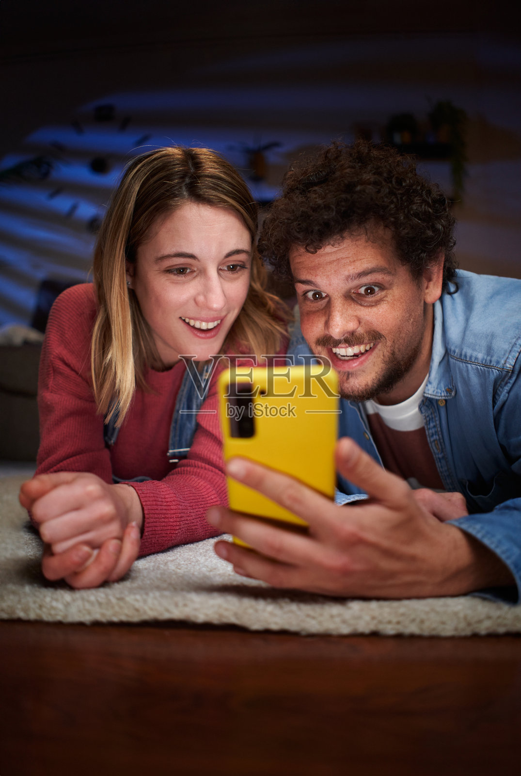 年轻的高加索情侣在夜晚的客厅里，惊讶地看着躺在地毯上的手机。照片摄影图片