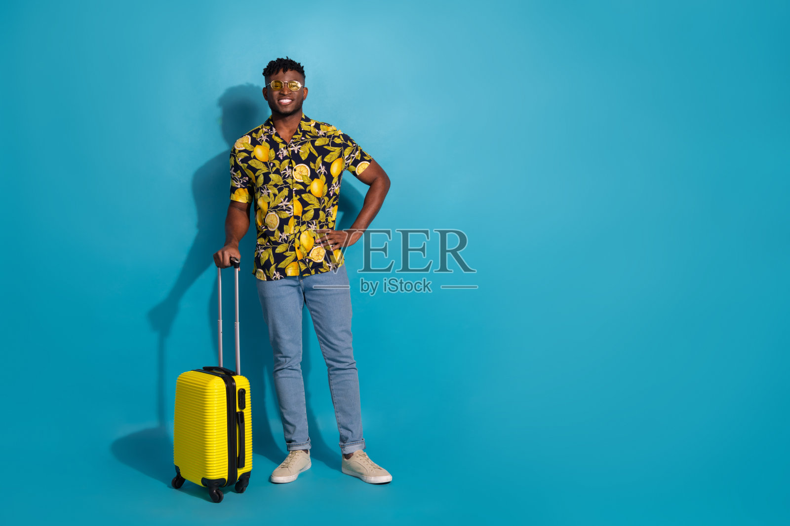 年轻男子穿着热带衬衫，手提黄色行李箱，背景为鲜艳的蓝色，自信地微笑。照片摄影图片