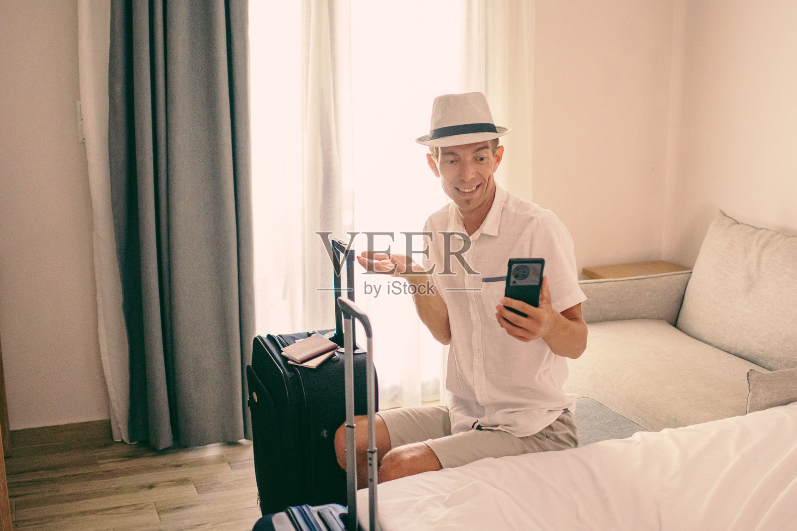 中年男子戴着草帽，手边放着打包好的行李箱和护照，坐在酒店房间里等待交通工具或预订他的航班，准备结束夏季假期。照片摄影图片