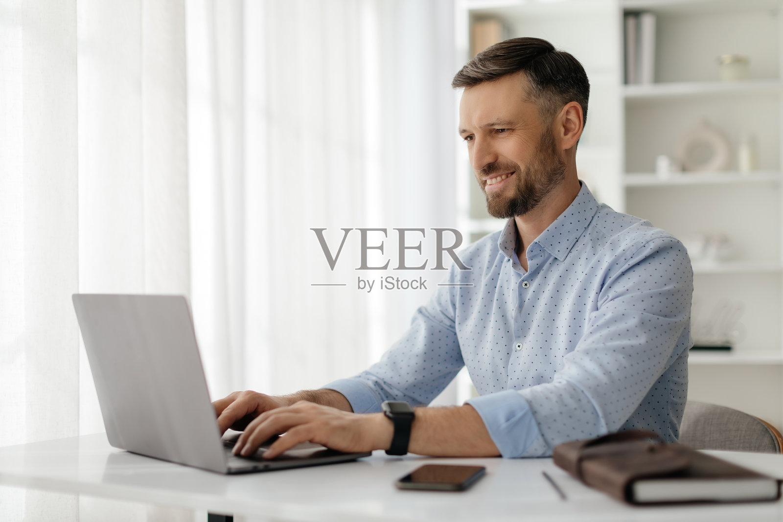 一名男子在明亮现代的家庭办公室里微笑着使用笔记本电脑，白天高效工作。照片摄影图片