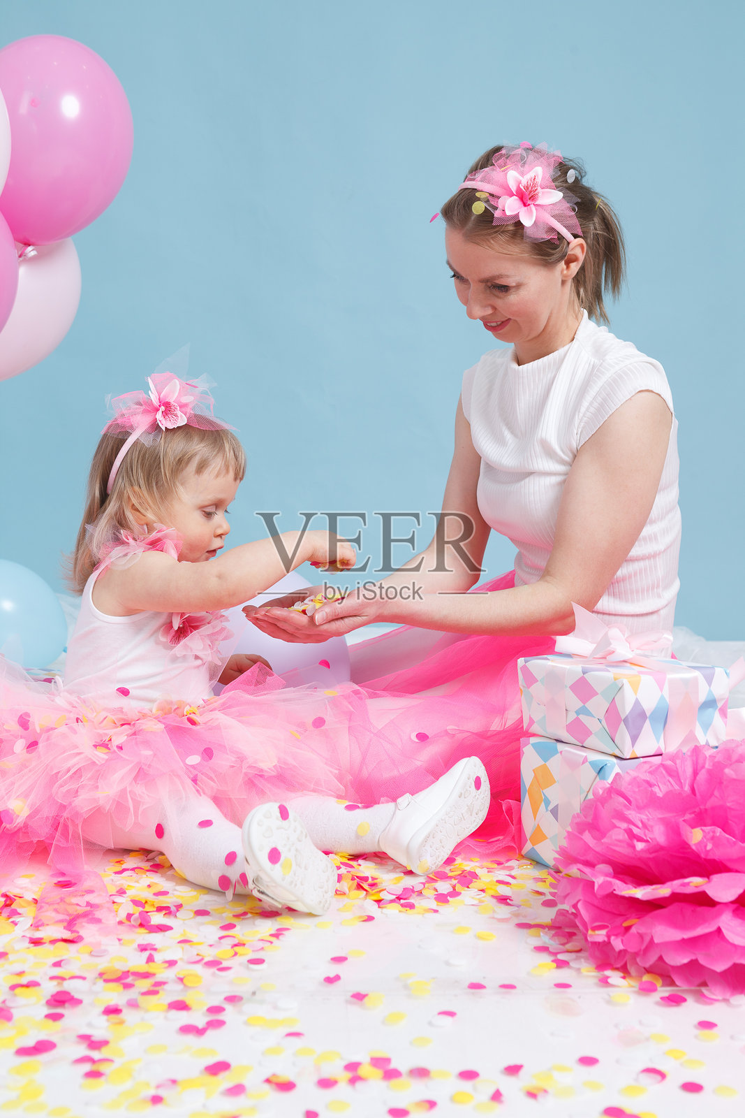 一个五彩纸屑的生日派对庆祝活动。一位母亲和她的小女儿穿着粉色裙子，坐在蓝色背景上。竖版图片。摄影棚拍摄。黄色、粉色、蓝色和白色。照片摄影图片