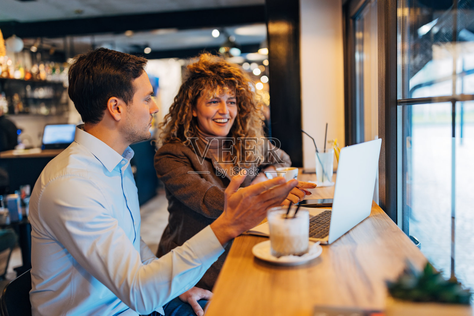 两位穿着休闲商务服装的同事在咖啡店远程工作。他们一边喝咖啡，一边使用笔记本电脑。照片摄影图片