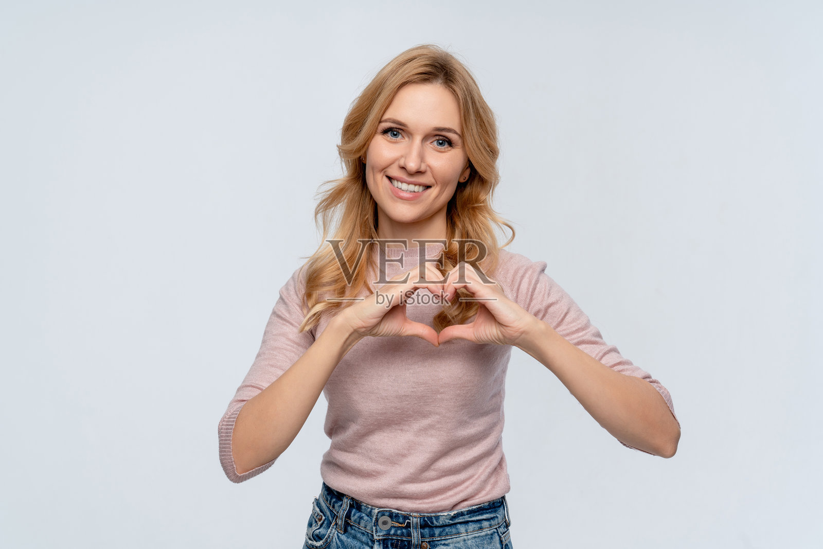 微笑的年轻女性穿着休闲服，站在白色背景前，满怀爱意地用双手做出心形符号。表达浪漫情感的情人节概念，日期为2月14日。照片摄影图片