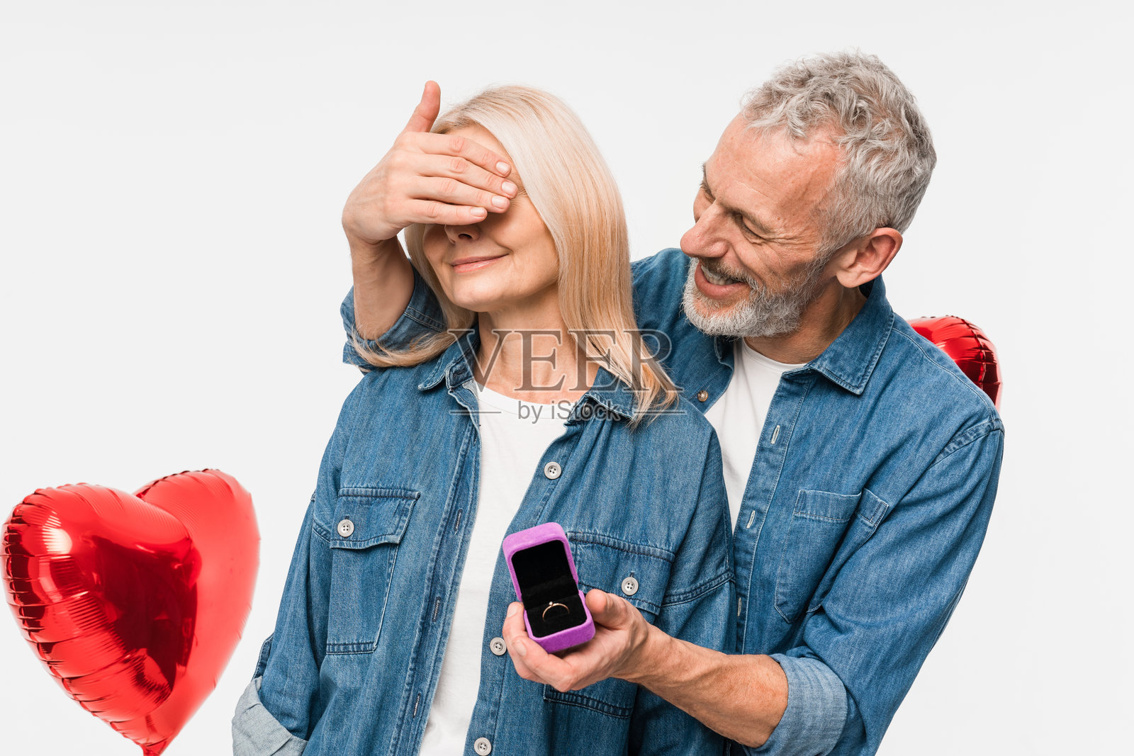 求婚。快乐的成熟男人在浪漫约会中向兴奋的女人展示打开的戒指盒，询问她是否愿意成为他的妻子，背景是红色心形气球，白色背景。情人节。照片摄影图片