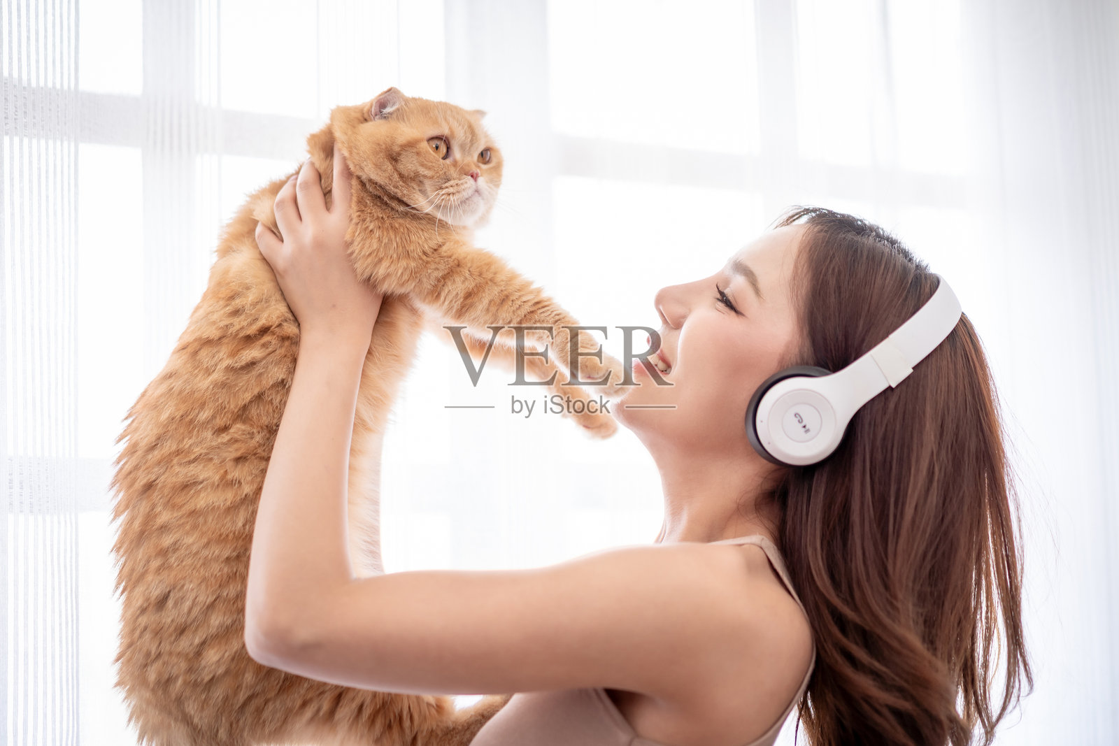 亚洲美丽的女性在家中幸福地抱着她的家猫。迷人的年轻女性宠物主人戴着白色耳机，悠闲地与她的小猫玩耍。照片摄影图片