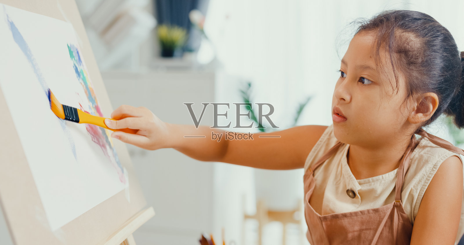 穿着围裙的亚洲幼女特写坐在沙发椅上，面前是画布和调色板，专注于在周末的家中用画笔创作抽象画。创意生活的孩子。照片摄影图片