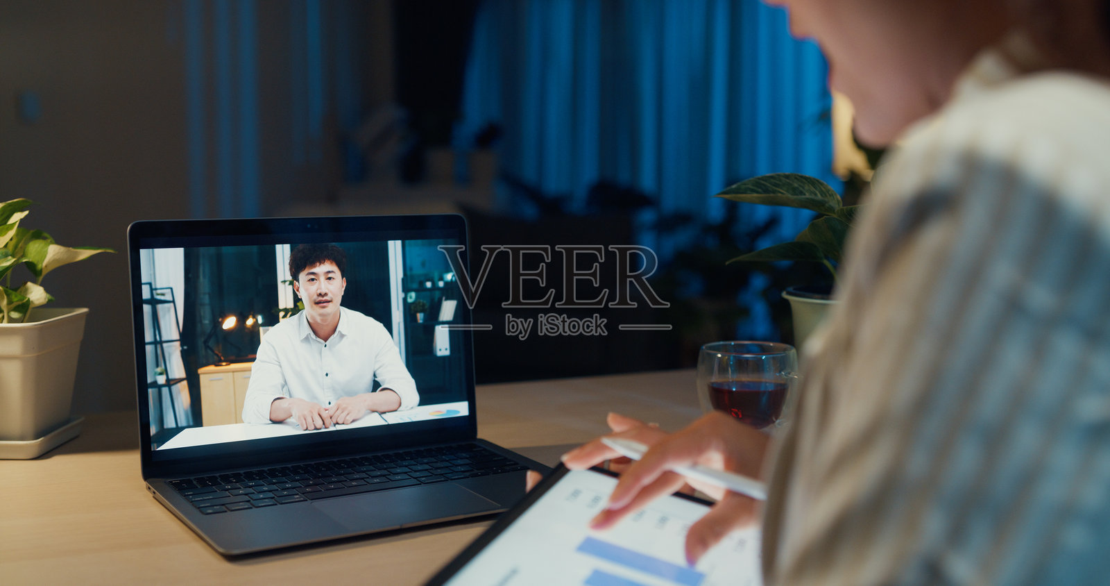 年轻的亚洲女商人穿着正式，坐在桌子前，手持平板和触控笔，专注于笔记本电脑屏幕，与同事进行在线会议，背景是温馨的家居客厅，夜晚加班工作。照片摄影图片