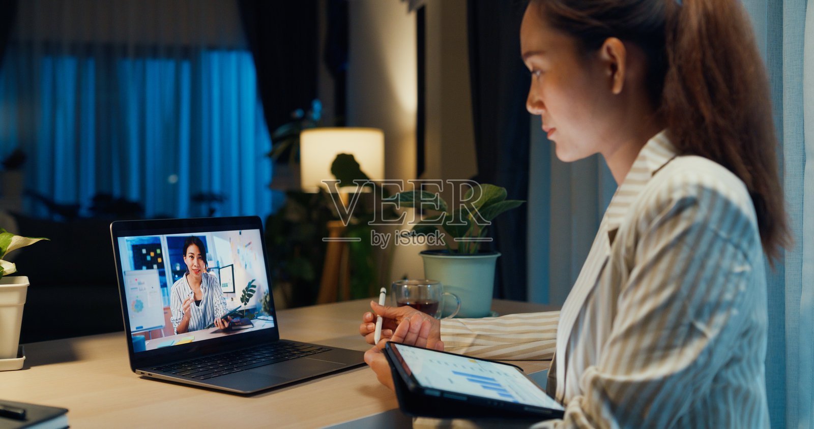 年轻的亚洲女商人穿着正式，坐在桌子前，手持平板和触控笔，专注于笔记本电脑屏幕，与同事进行在线会议，背景是温馨的家居客厅，夜晚加班工作。照片摄影图片