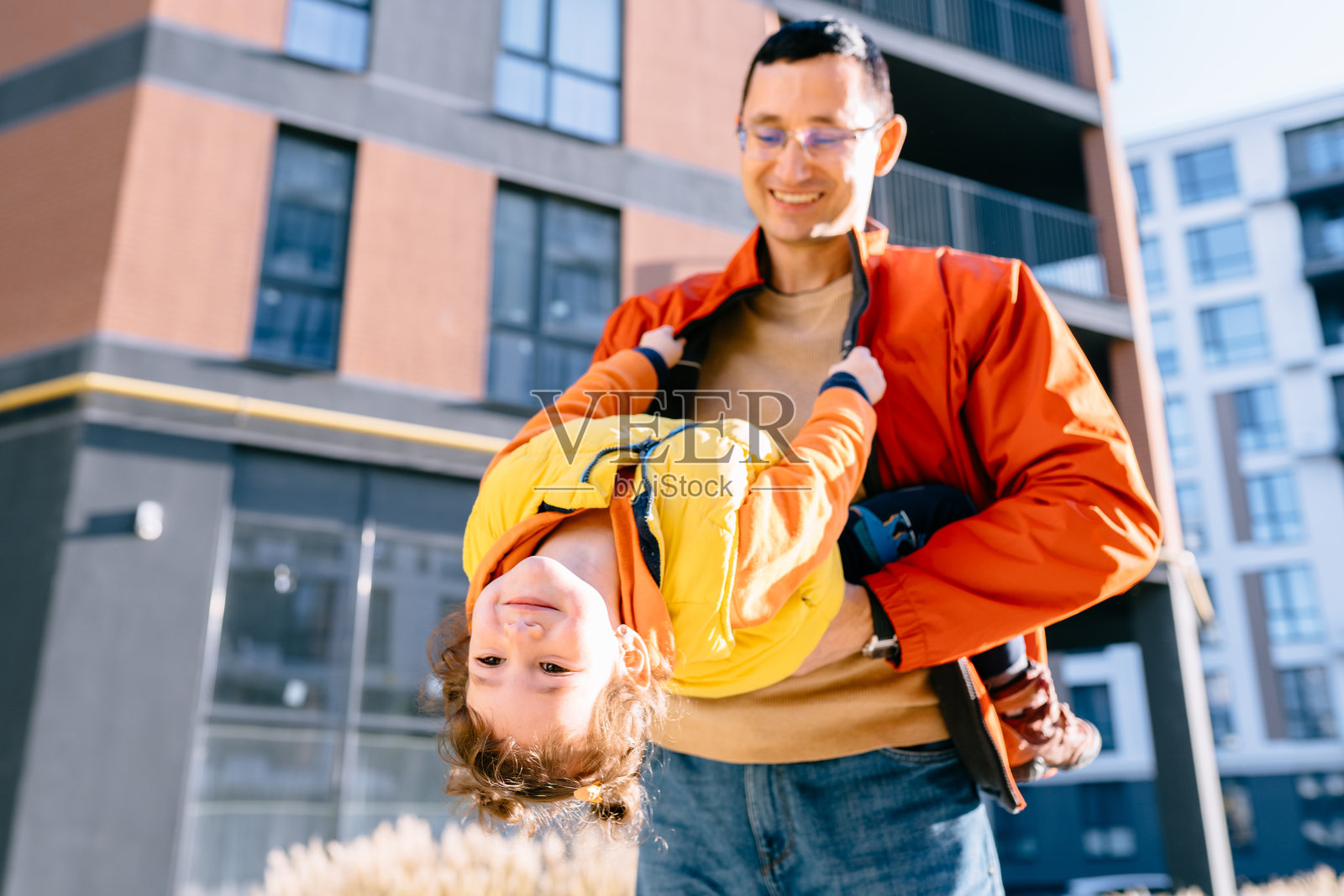 活泼的家庭活动。快乐的父亲在现代城市建筑附近把女儿倒提着。照片摄影图片