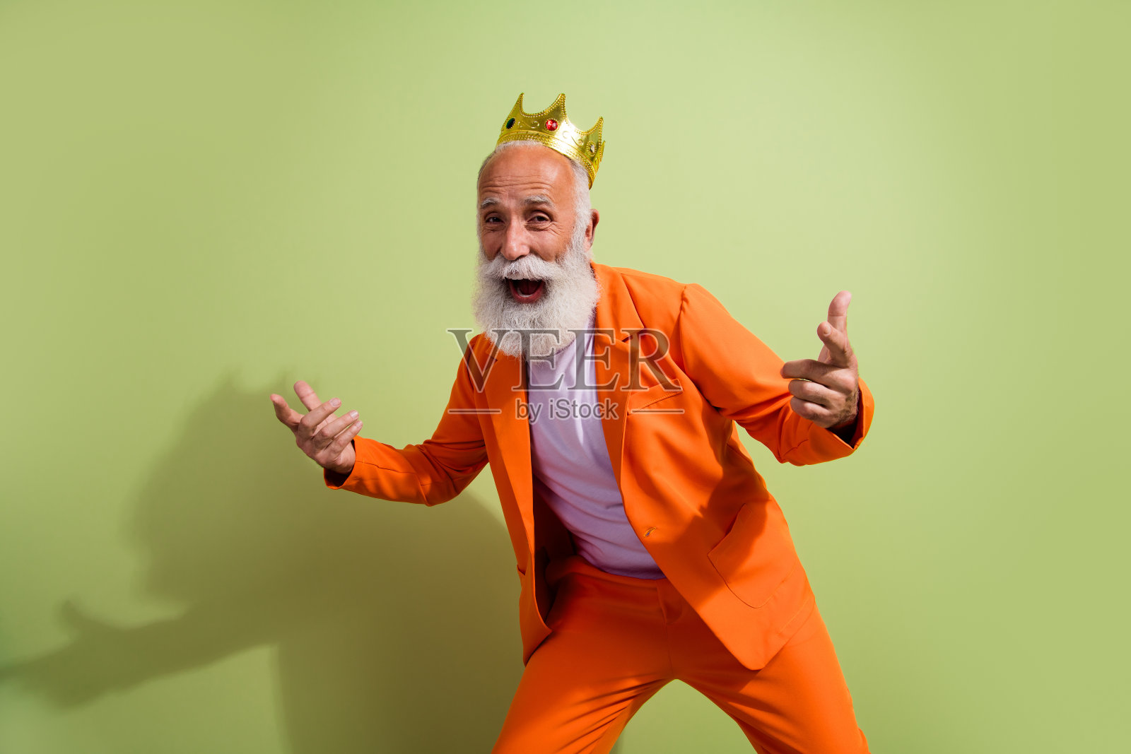 穿着橙色西装、戴着王冠的老年男演员在绿色背景下欢欣鼓舞。照片摄影图片