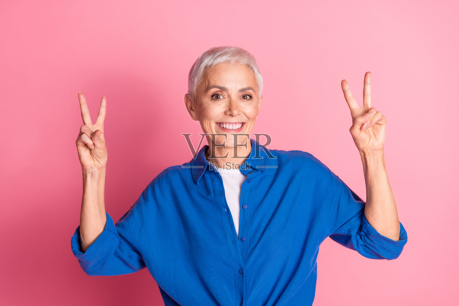 迷人积极的女性穿着蓝色衬衫，双手比出两个V字手势，背景为空旷的蓝色。照片摄影图片