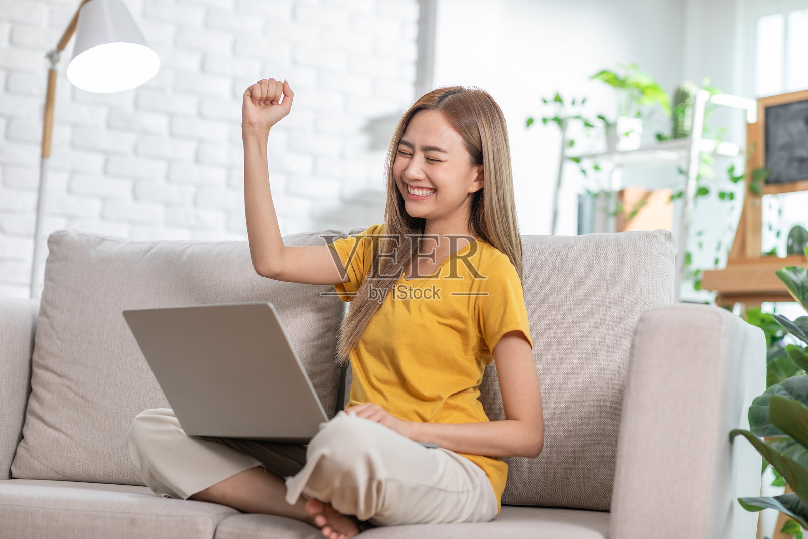 年轻女子穿着黄色衬衫，坐在舒适的沙发上，举起拳头庆祝，目光注视着她的笔记本电脑。照片摄影图片