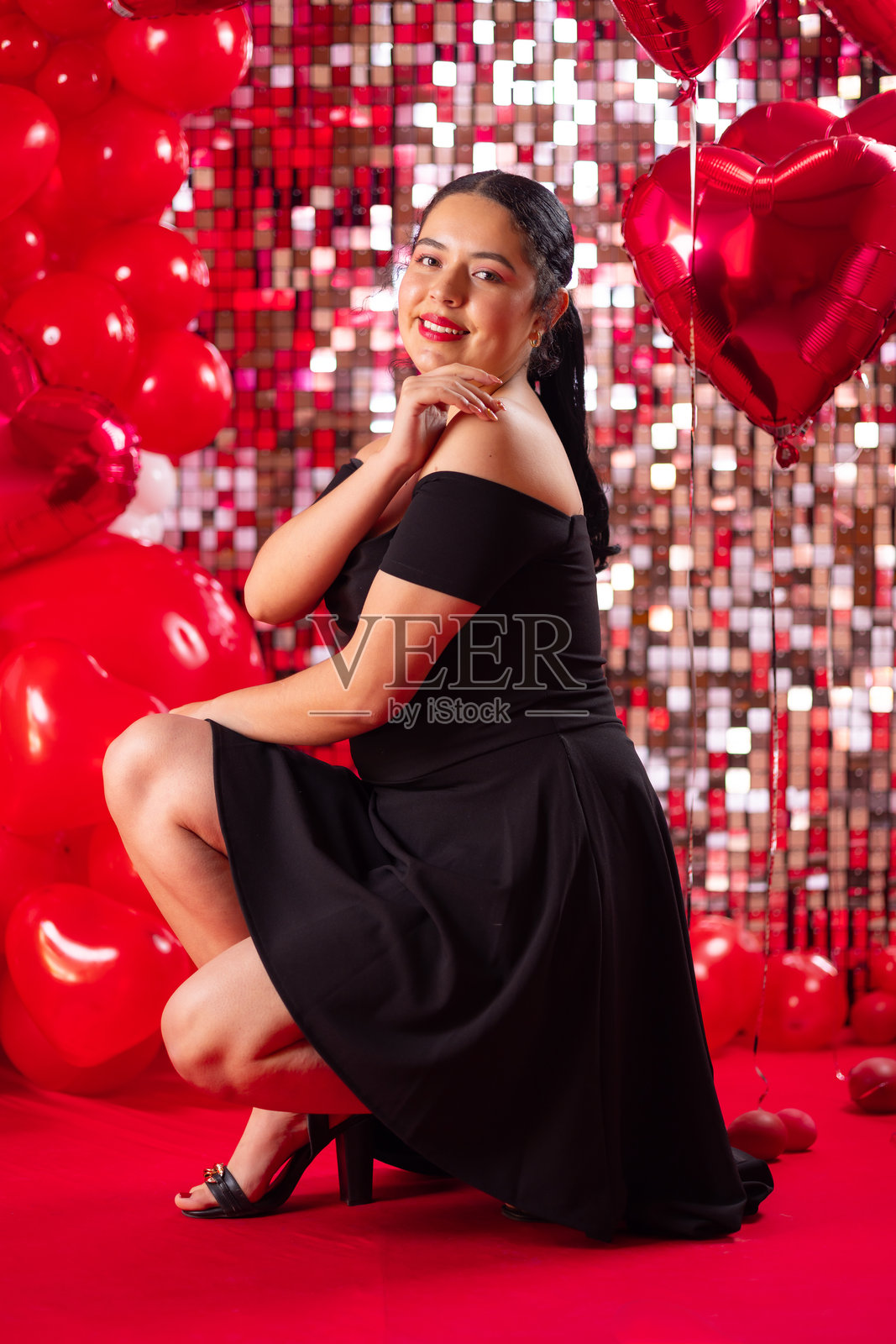 身穿黑色连衣裙的美丽年轻女性在红色调的心形气球背景前摆姿势。照片摄影图片
