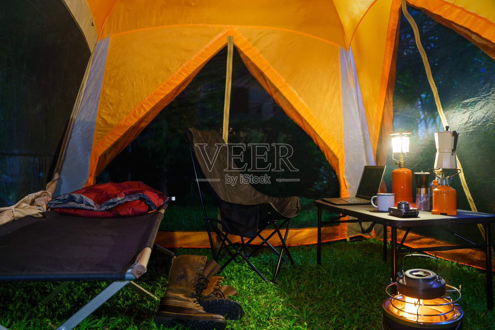 森林夜晚的露营帐篷内有折叠床、煤气灯和咖啡套装，木质露营桌上放着笔记本，以及户外创意。照片摄影图片