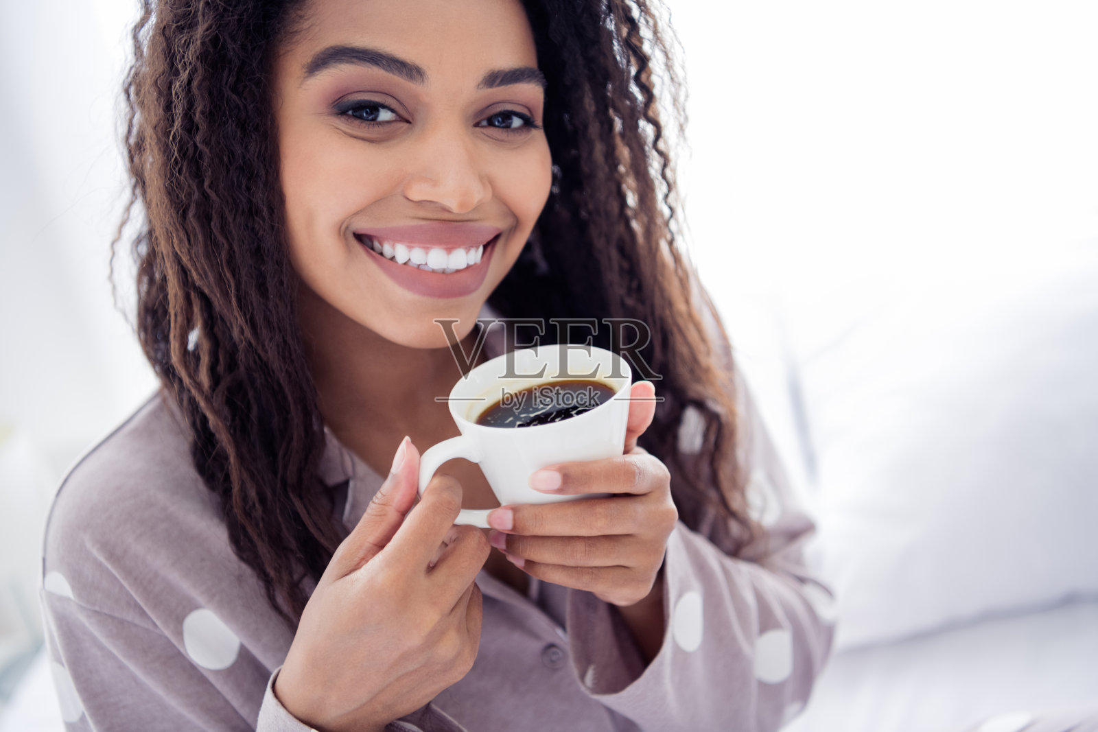 年轻女性在床上享受早晨咖啡，脸上洋溢着幸福的微笑，阳光透过窗户洒入房间。照片摄影图片