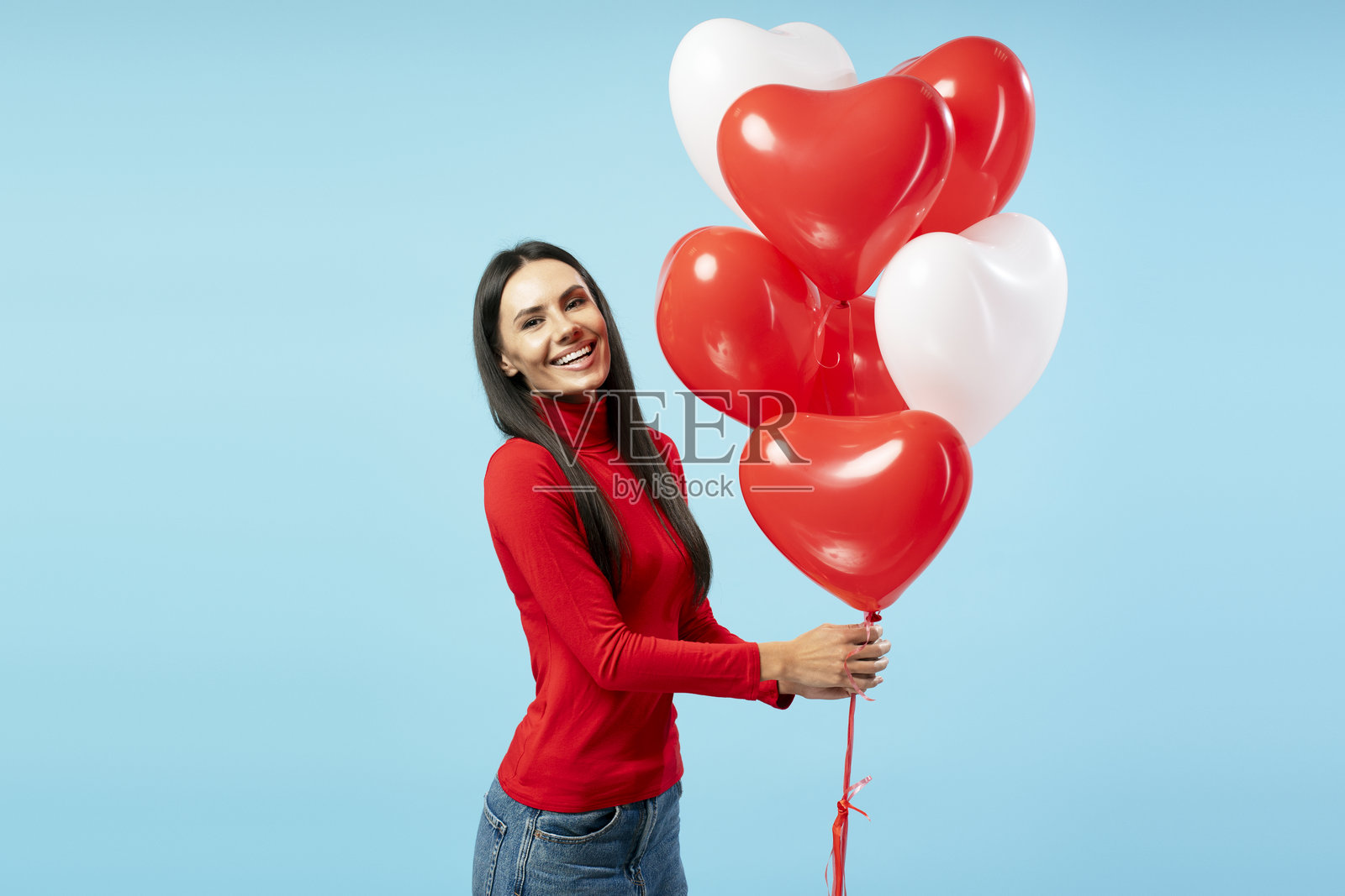 快乐的中年拉丁裔女性手持红白相间的心形气球，背景为蓝色。照片摄影图片