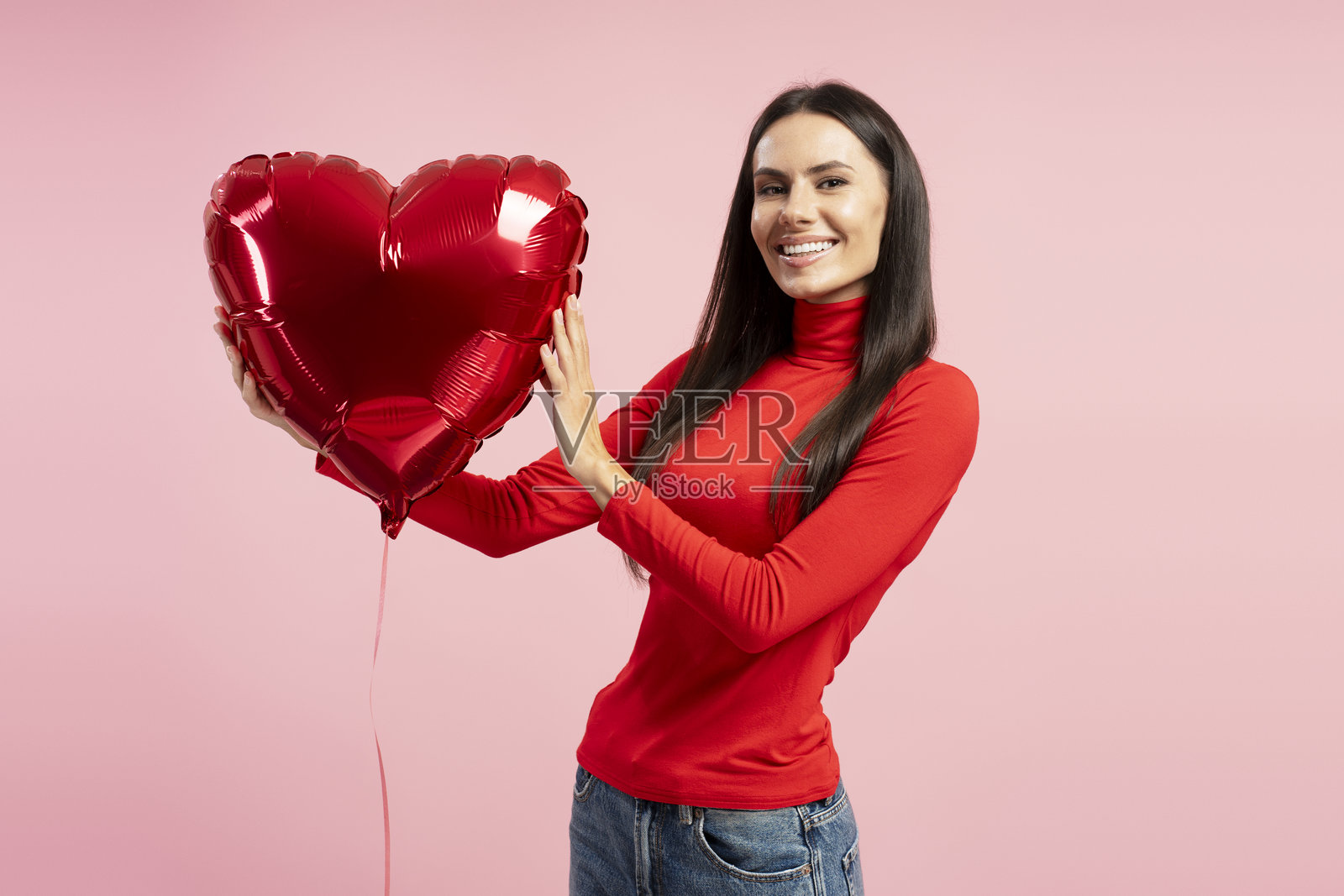美丽的女人在粉色背景上微笑，手中拿着红色心形气球照片摄影图片