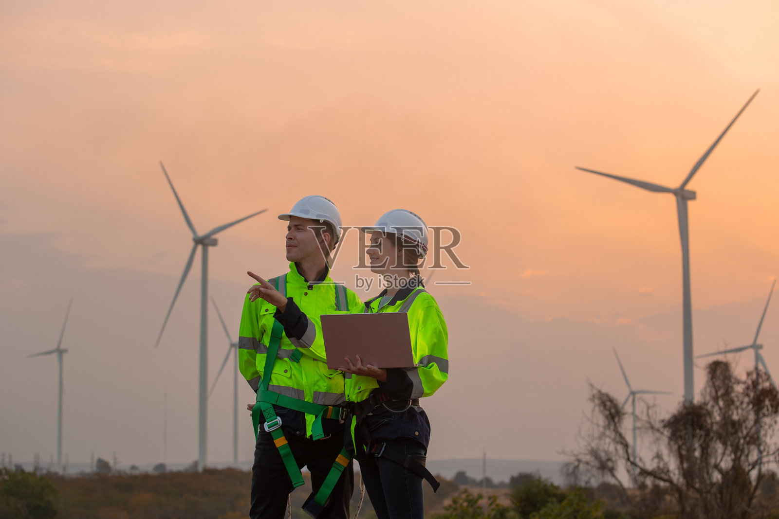 穿着安全制服的工程师团队在夕阳下的风能发电站讨论可再生能源计划，手中拿着笔记本电脑。科技环境有助于减少全球变暖问题。照片摄影图片