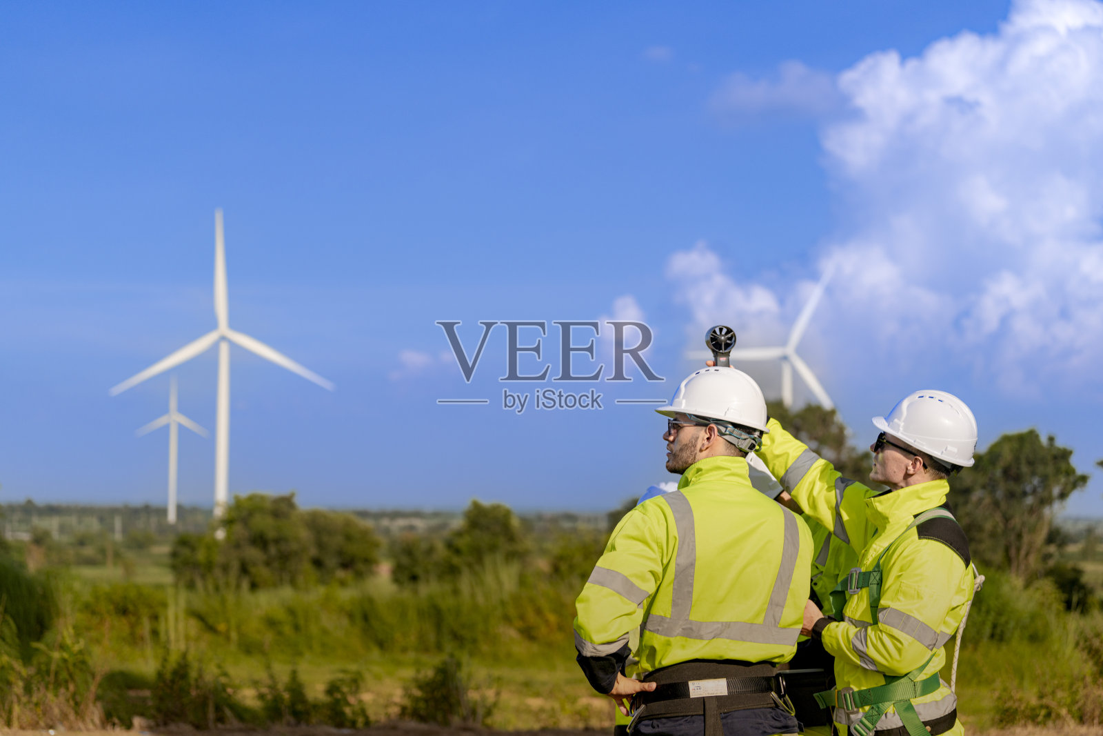 一组穿着高可见度安全装备的工程师在风电场聚在一起，进行检查并讨论可再生能源项目。周围是风力涡轮机，天空晴朗。照片摄影图片