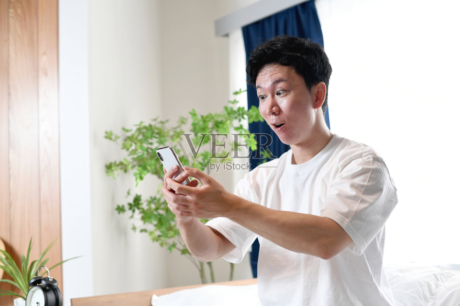 惊讶的亚洲男子坐在床上，手里拿着智能手机照片摄影图片