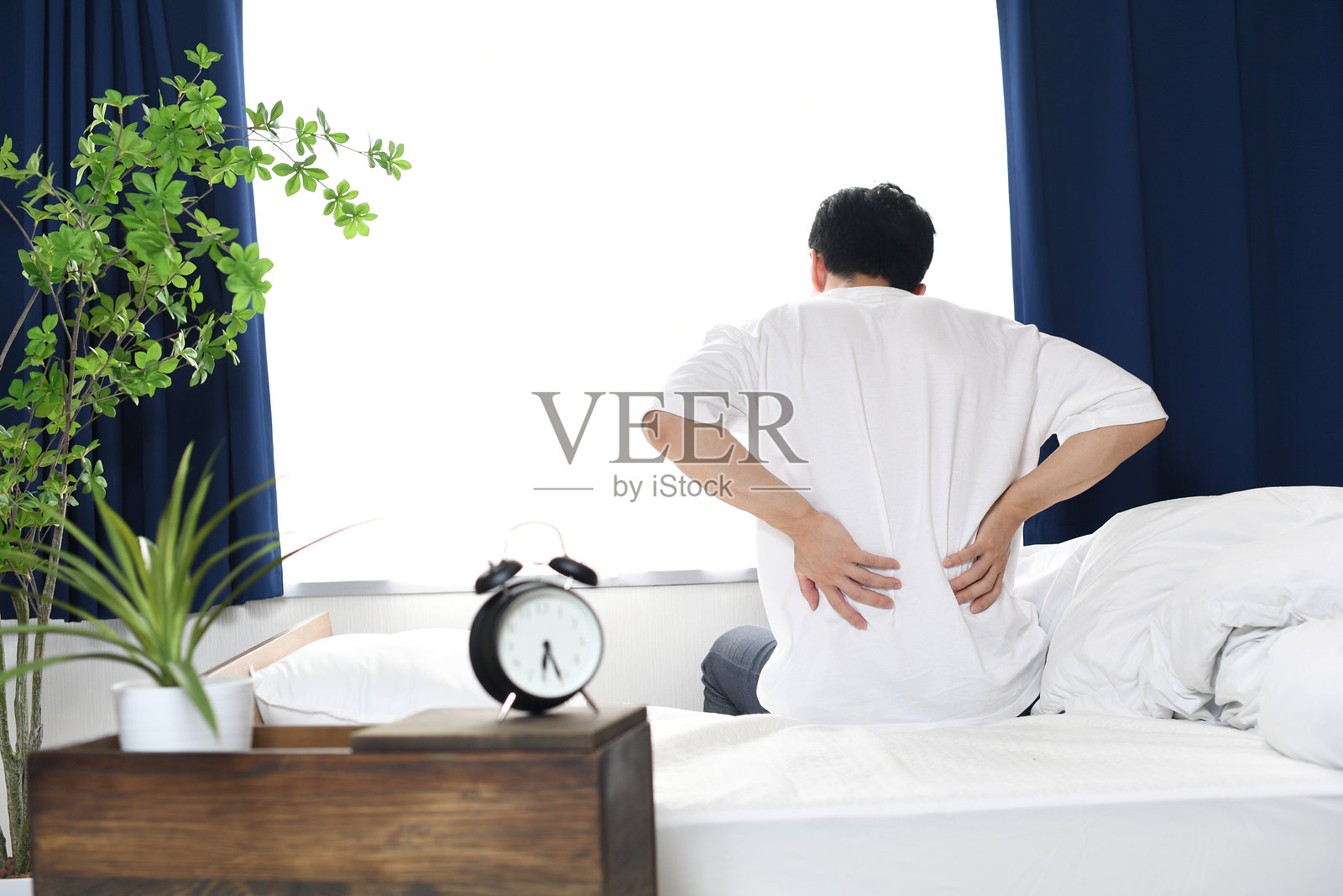 年轻男子坐在床上，双手抱着腰，背影。照片摄影图片