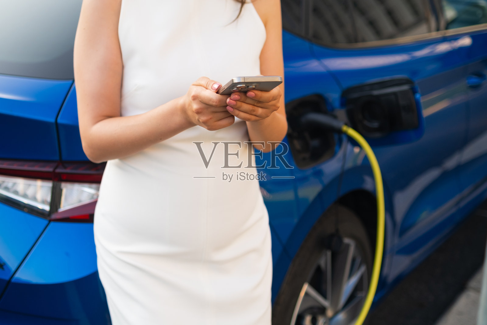 女性在阳光明媚的日子里一边使用智能手机一边为蓝色电动车充电。照片摄影图片