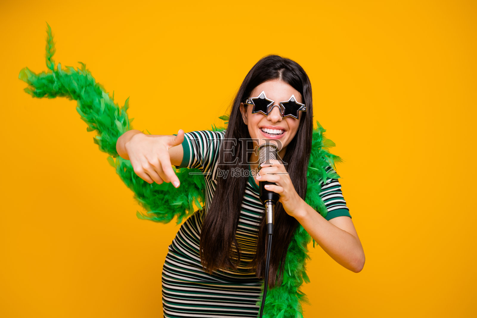 明亮的肖像，一位快乐的年轻女性佩戴着星形太阳镜和绿色羽毛围巾，背景为鲜艳的黄色。照片摄影图片