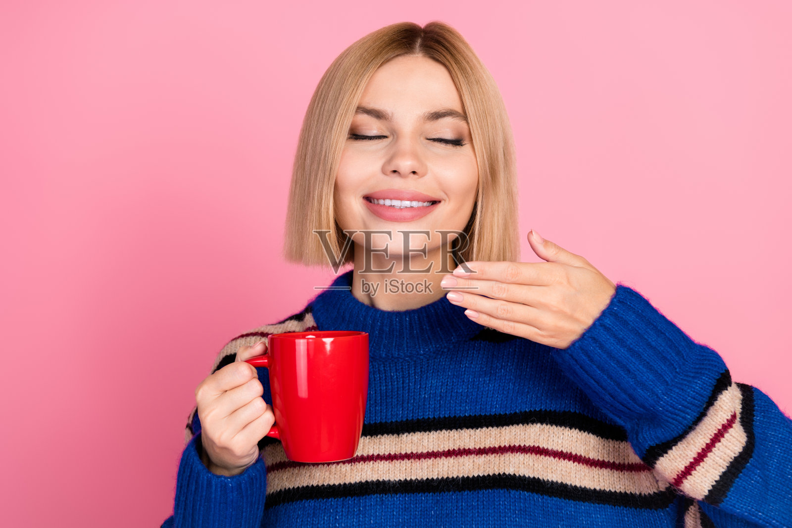 一位年轻女性穿着彩色条纹毛衣，享受着一杯红色饮品，背景为粉色。照片摄影图片