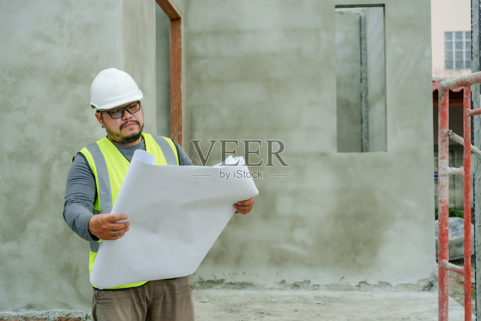 工人在户外建筑工地上，穿着白色安全头盔和亮黄色反光背心，手持大型蓝图，进行检查和仔细规划，周围有脚手架和混凝土墙。照片摄影图片