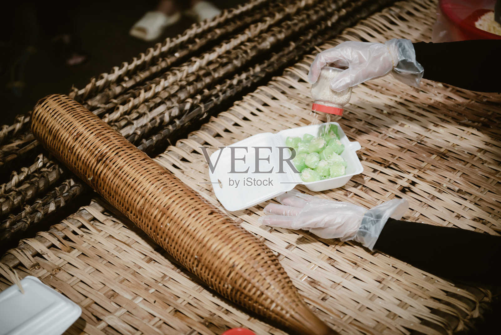 亚洲女性戴着一次性食品手套，手工为传统南越绿米包糕Banh Hu Tieu Ngot添加芝麻，放在泡沫容器中，背景是竹席，旅行于芹苴。照片摄影图片