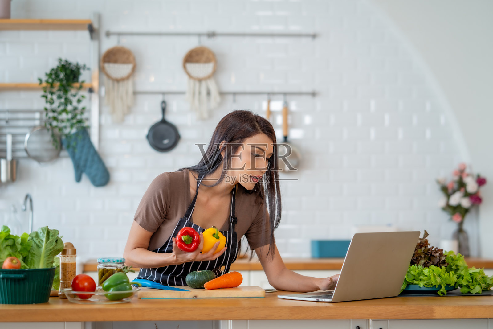 女性在厨房准备健康食品，同时使用笔记本电脑。她手里拿着五彩缤纷的蔬菜，展示现代烹饪环境。照片摄影图片