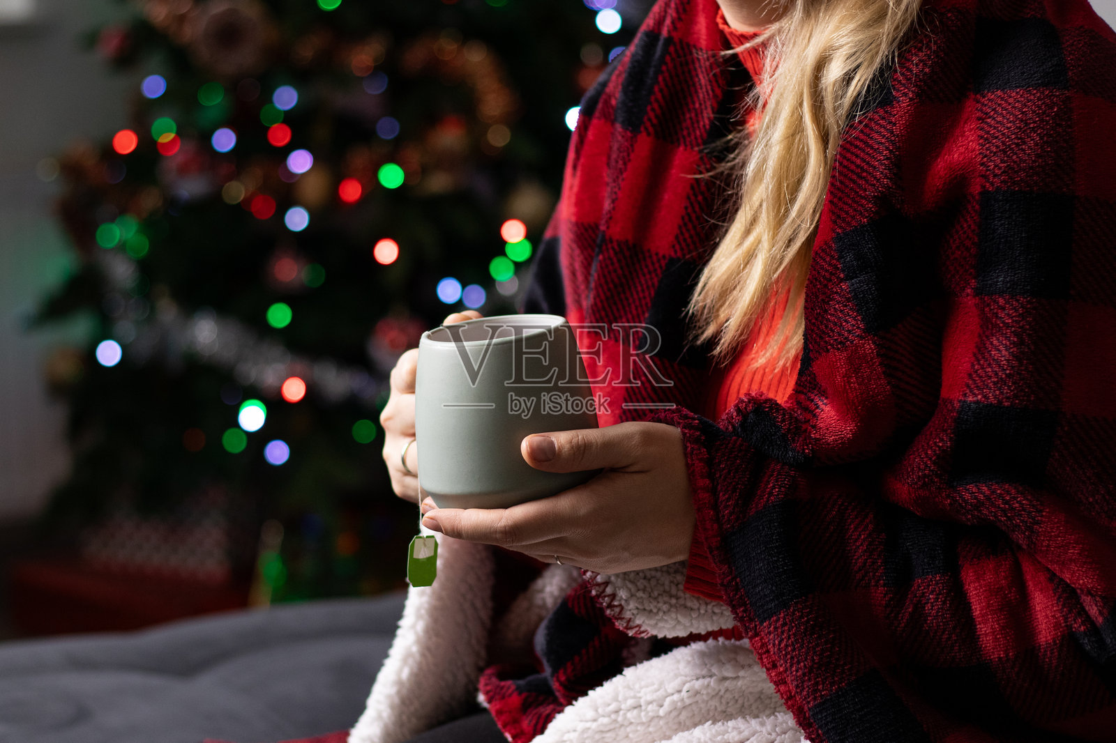 特写镜头：一位女性手握绿色杯子，杯中热饮，坐在沙发上，身披红色毛毯。照片摄影图片