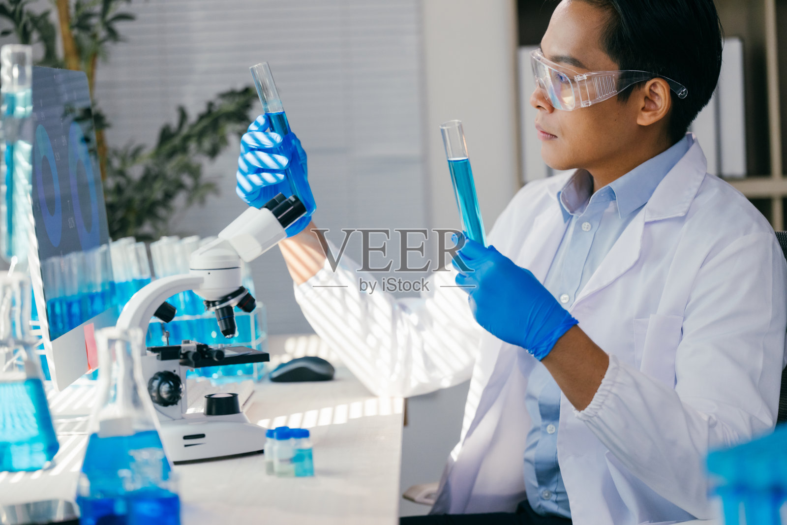 年轻科学家穿着实验室外套和手套，比较两个装有蓝色液体试剂的试管，在现代研究实验室中进行实验，旁边有显微镜和电脑。照片摄影图片